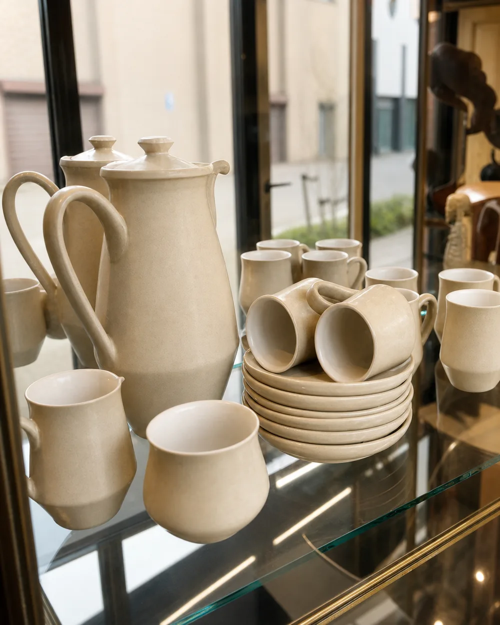 Roomkleurig steengoed koffieservies met kannen, kopjes en schotels in een glazen vitrine.