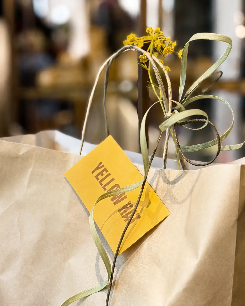 Papieren draagtas met een geel Yellow Hat-label, raffia lint en een takje gele bloemen.